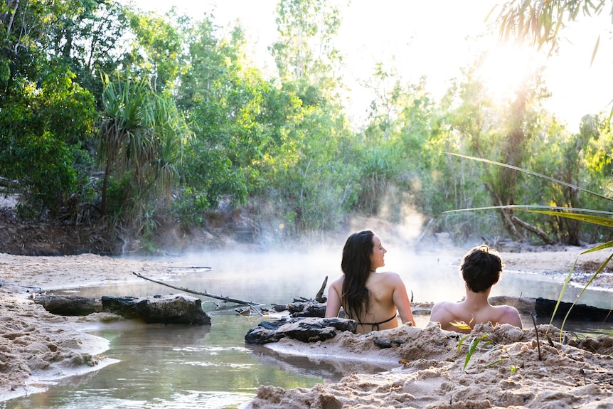 Two people sit in hot springs water surrounded by steam and sand