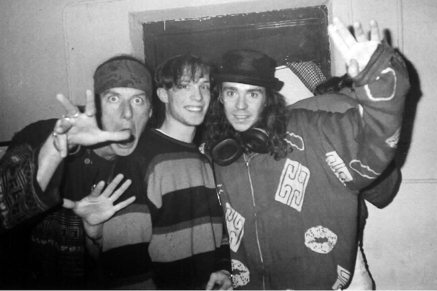 Three young men act up for the camera, black and white photo, man on right wears top hat and has large headphones around neck.