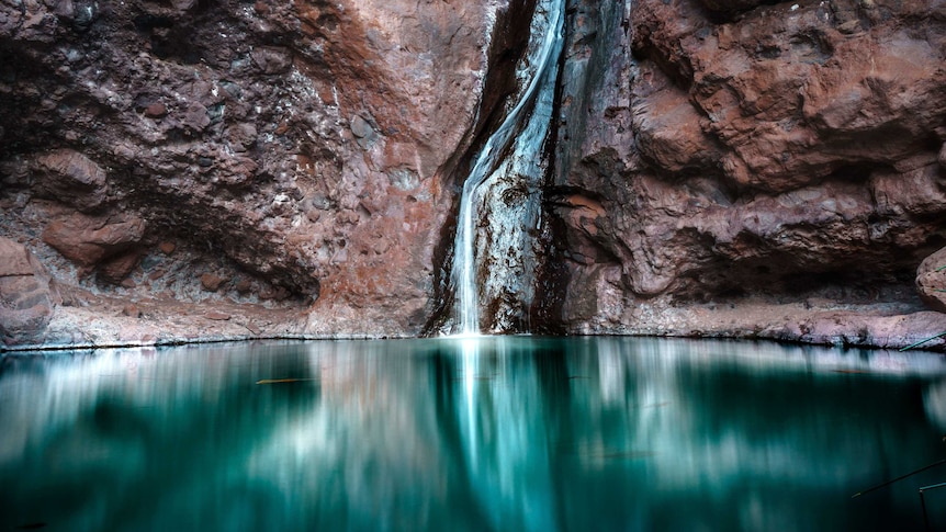A wall of reddish rock with a stream of water in its middle, leading to a deep, aqua-coloured pool of shiny water.