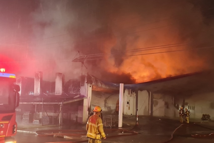 A firefighter stands before a fire sweeping across the roof of a commercial building in the early morning.