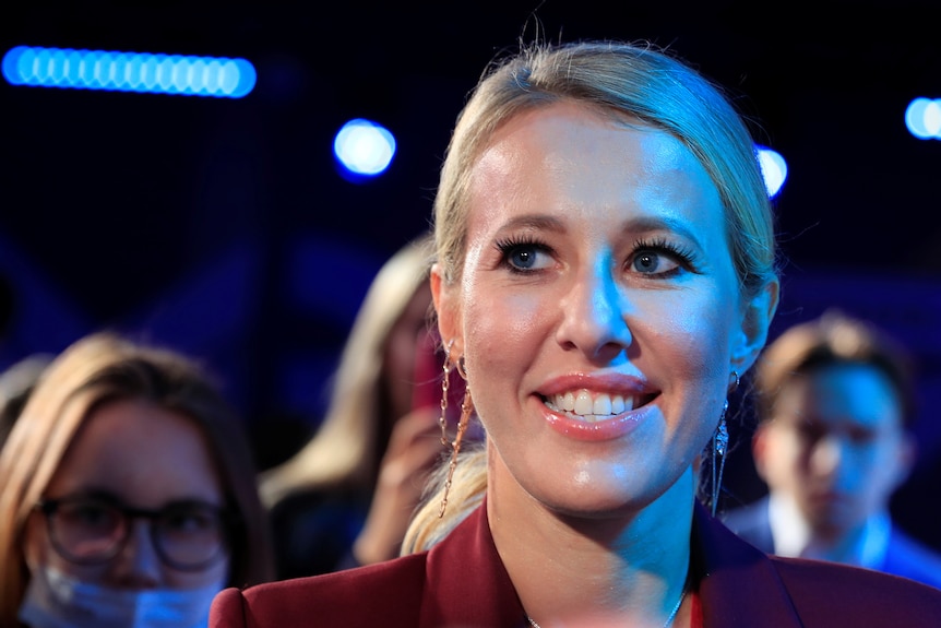 Ksenia Sobchak, wearing a burgundy blazer and dangly earrings, her blonde hair in a ponytail smiles from a crowd