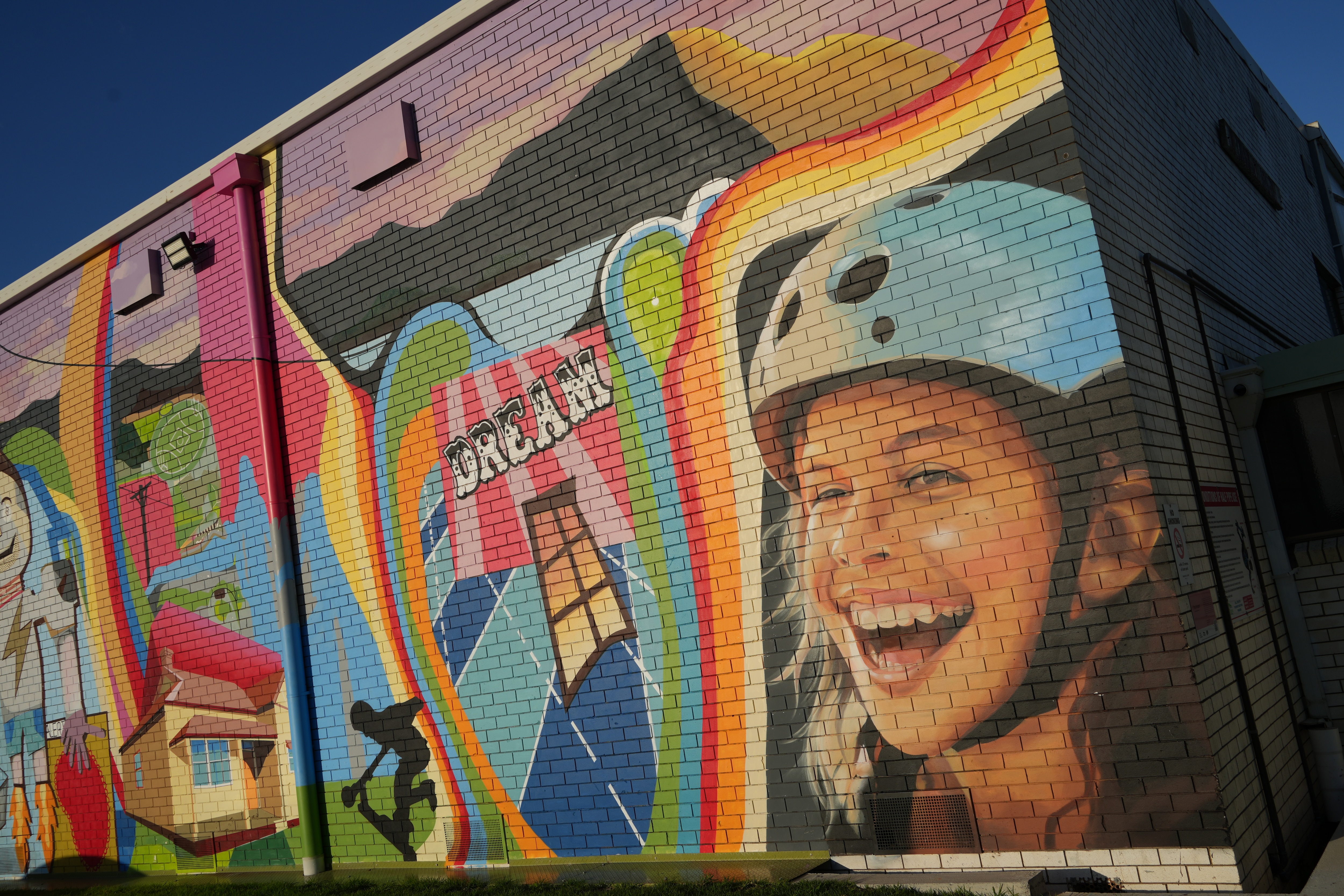 Colourful mural on a brick wall next to skate park, showing girl smiling wide wearing a blue helmet.