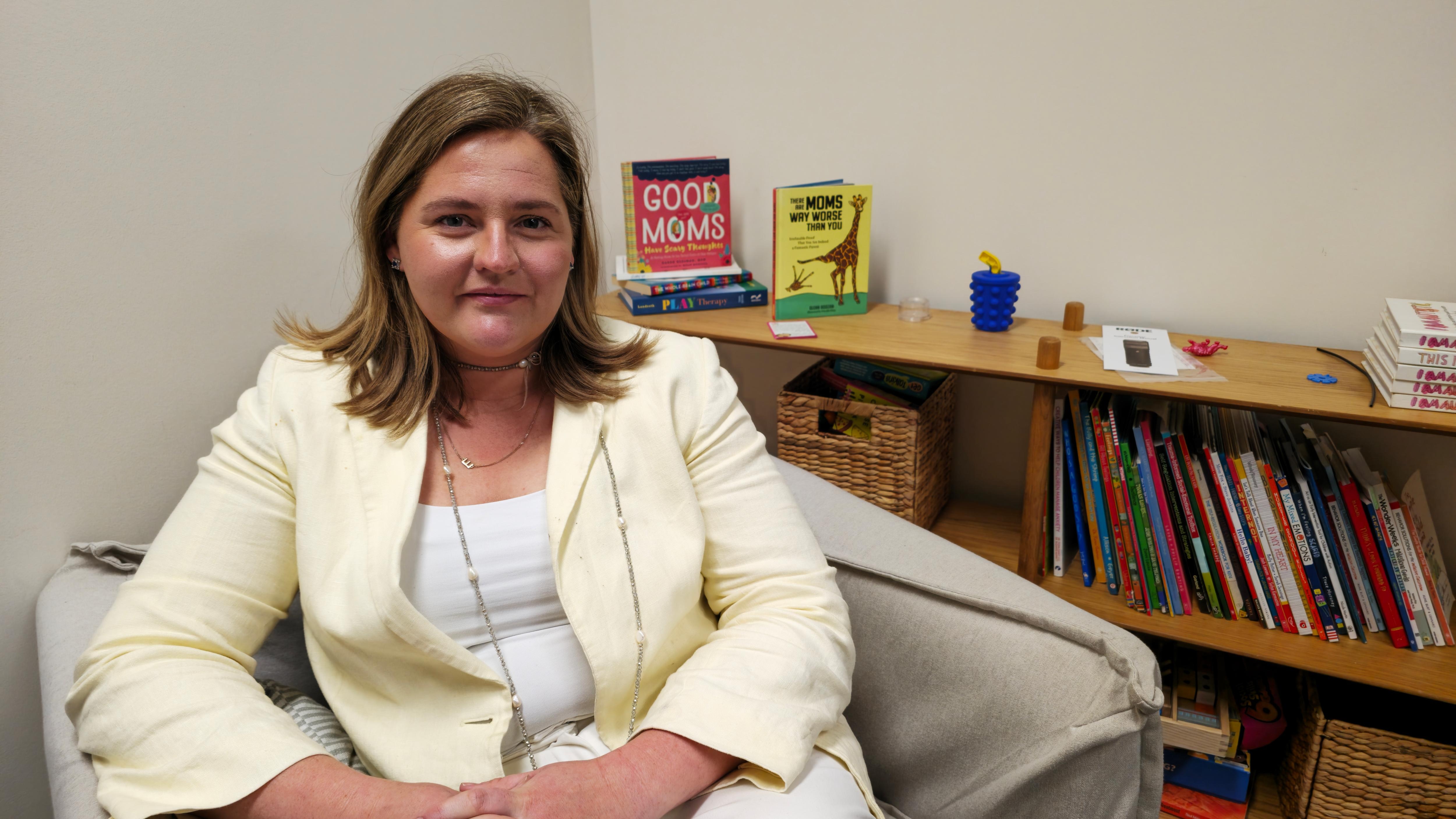 A woman (Jen Laurie) with shoulder-length hair sits in an armchair in front of a bookcase.