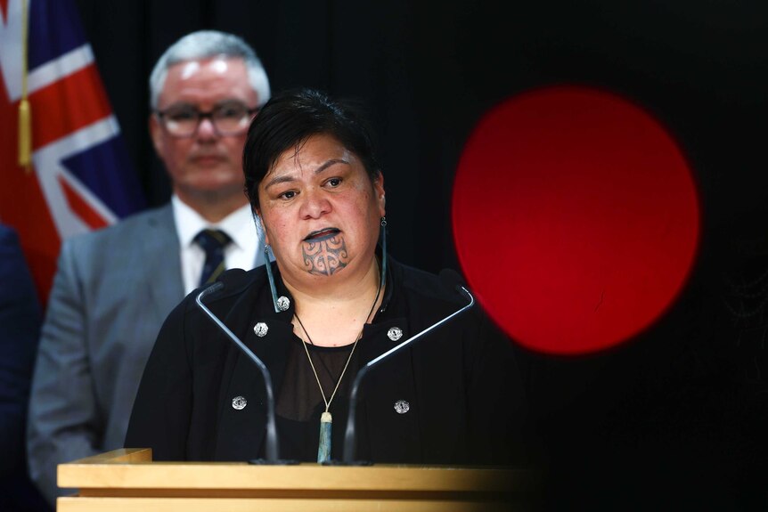 A Maori woman with traditional chin and lip tattoos speaks at a podium.