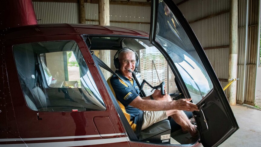 A mid shot of a man sitting in a maroon helicopter with the door open in a shed