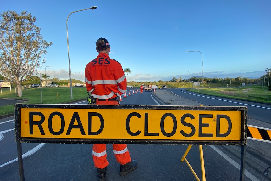 A man in an SES uniform stands with his back facing a 'road closed' sign