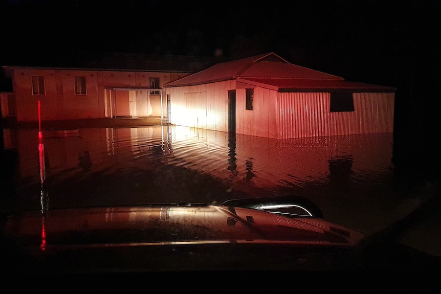 A nighttime photo of a corrugated iron hall surrounded by floodwaters, illuminated by car headlights.