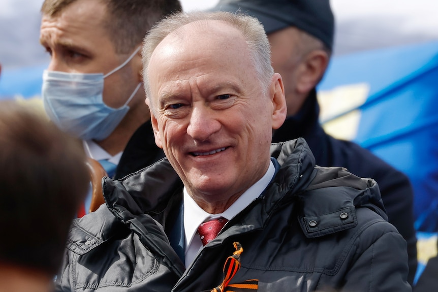 An older man with graying hair smiles while wearing a puffer coat over his suit and red tie