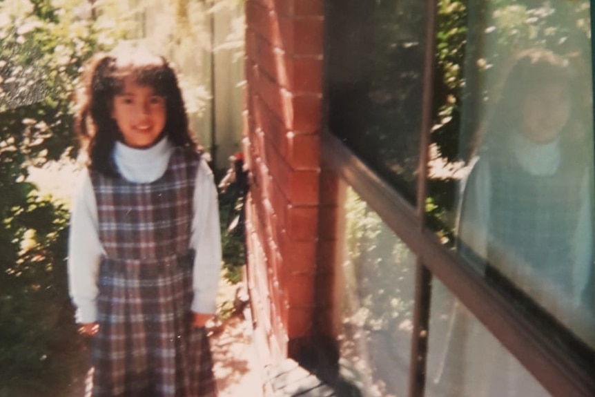 A photograph of a girl in a school dress standing next to a brick house, smiling.