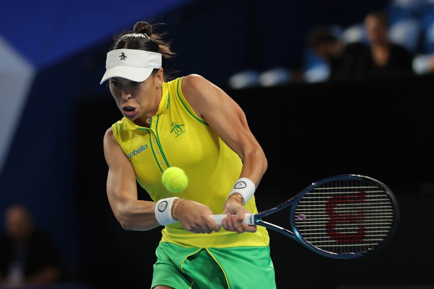 A woman wearing yellow and green swings her tennis racquet to hit a ball.