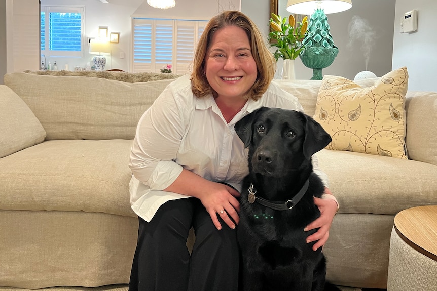 Jaci Armstrong sits on her lounge in her living room hugging her black guide dog