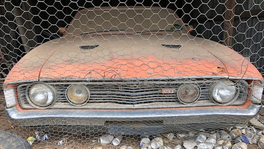 An orange car covered in dirt sits in a shed behind wire.
