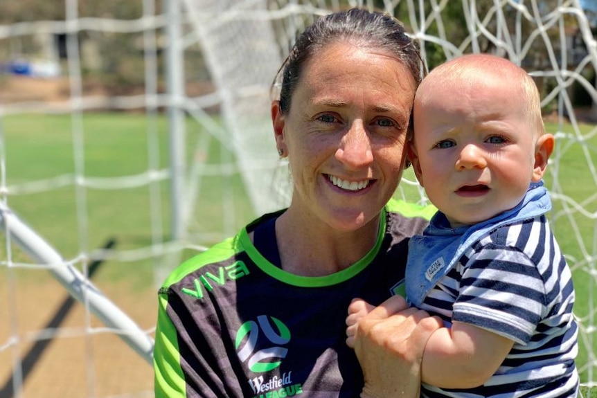 A woman holds her child in her arms as they stand in front of a soccer net.