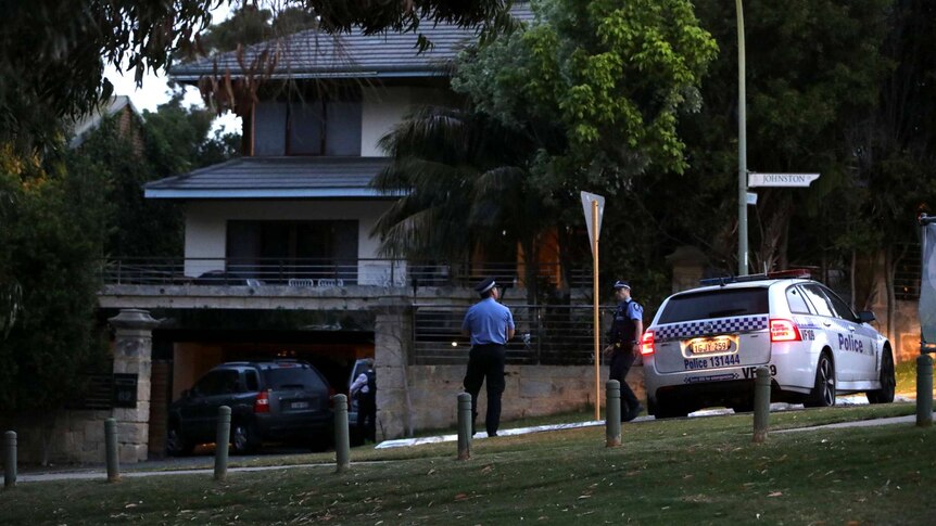 Two police officers stand next to a police car outside a large house in Mosman Park.