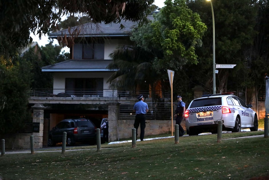 Two police officers stand next to a police car outside a large house in Mosman Park.