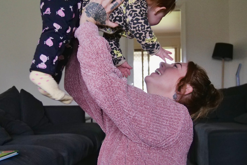 A laughing woman, red hair tied up, pink jumper, hoists baby up, both look away from camera, floor lamp, lounge in background.