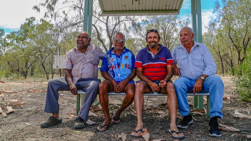 Four men sit on a bench in front bushland.