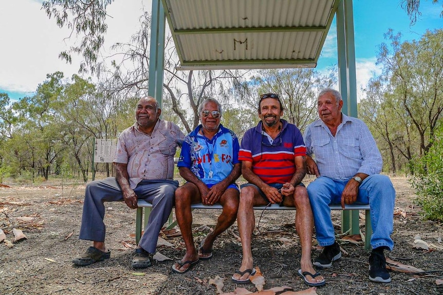 Four men sit on a bench in front bushland.