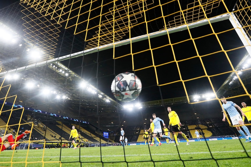 The ball (with the Champions League logo on it) hits the net for a goal, with players in the background. 
