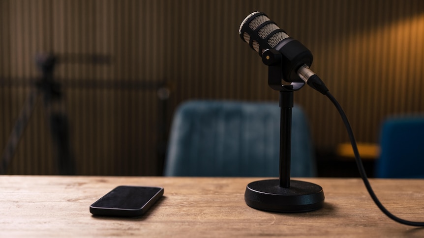 Image of a microphone sitting on a desk next to a smart phone. The chair in front of the desk is empty.