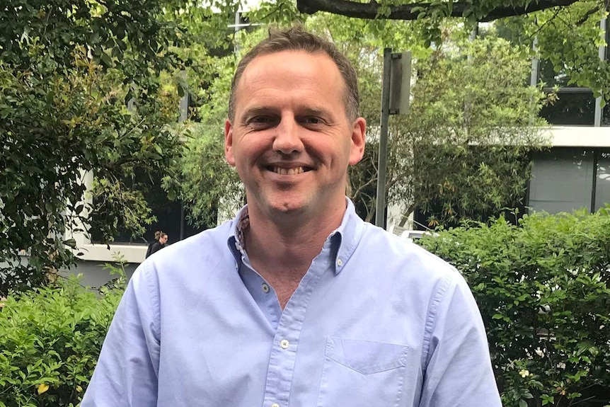 A man smiles while wearing a blue shirt with green trees in the background.