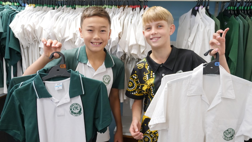 Two boys holding up uniform polo shirts with a bunch of shirts on a rack behind them. 