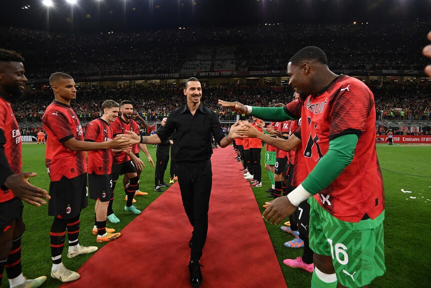 Soccer star Zlatan Ibrahimovic smiles and clasps hands with his teammates as he walks down a red carpet on the pitch.