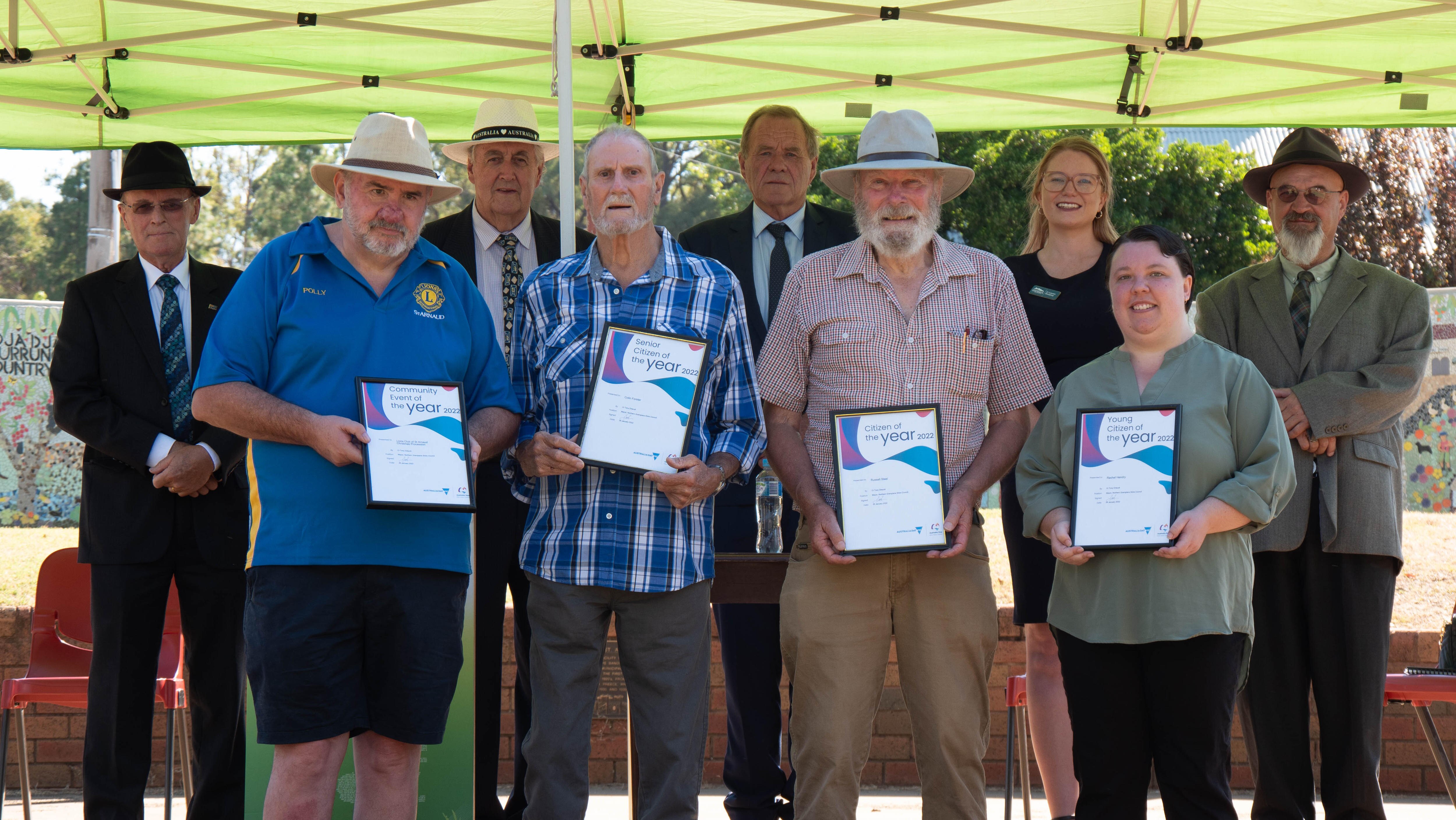4 people, two with hats, one with a blue plaid shirt hold their framed awards, while 4 men and woman (councillors) stand behind