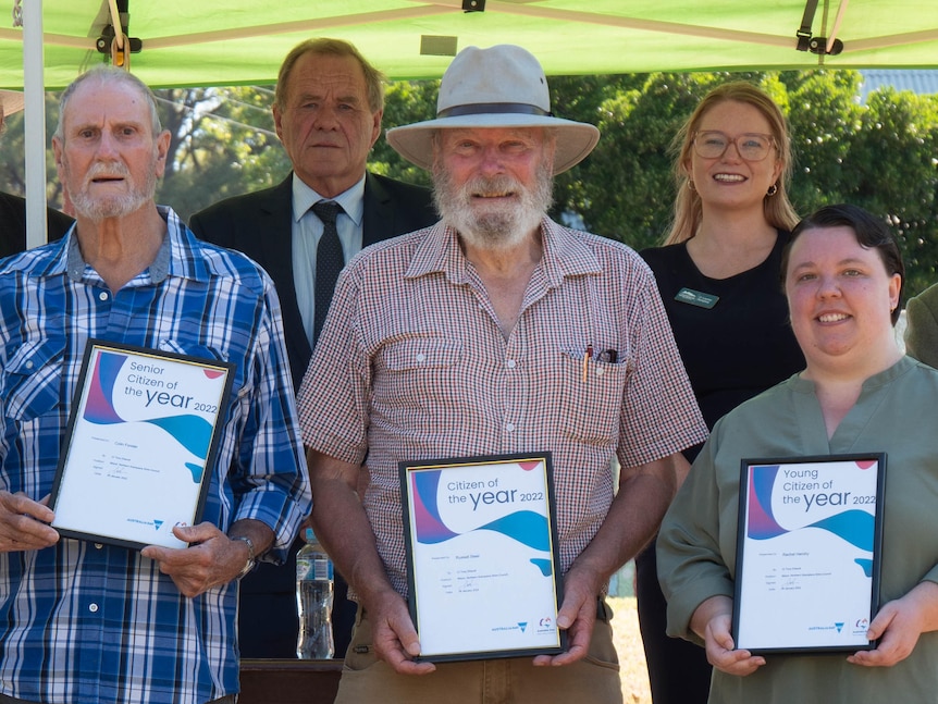 4 people, two with hats, one with a blue plaid shirt hold their framed awards, while 4 men and woman (councillors) stand behind