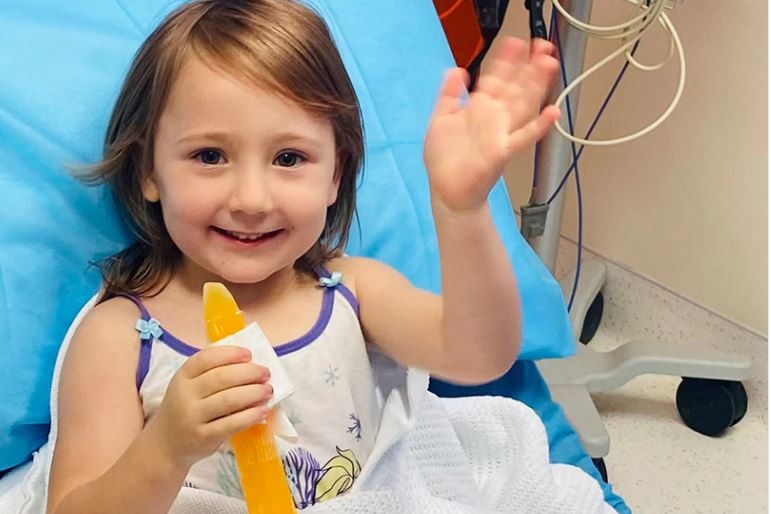 A girl with short orange hair waves from a hospital bed