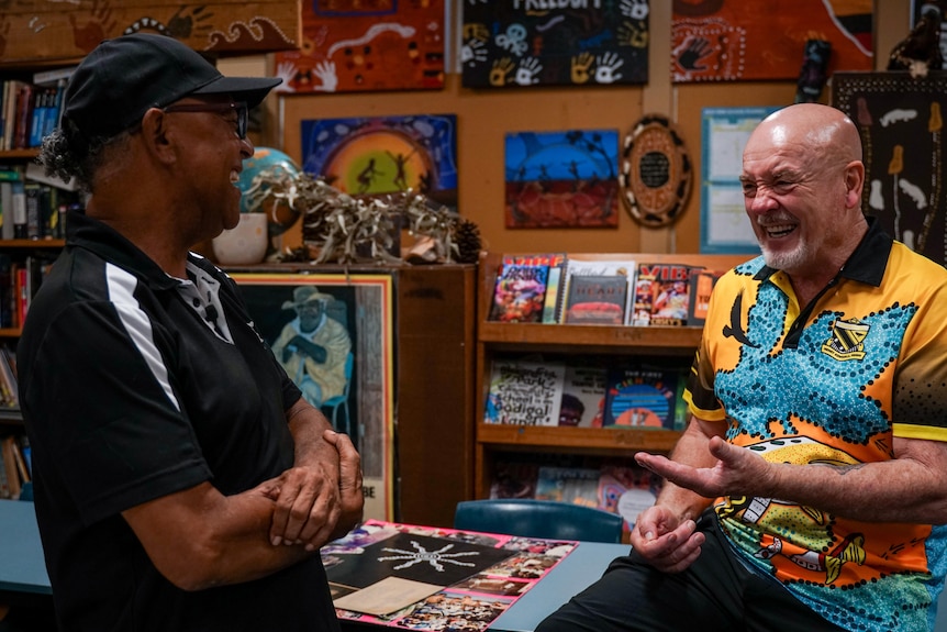 Two men, one wearing black t-shirt and cap, the other bald, a yellow and blue tee in a room with Indigenous paintings.