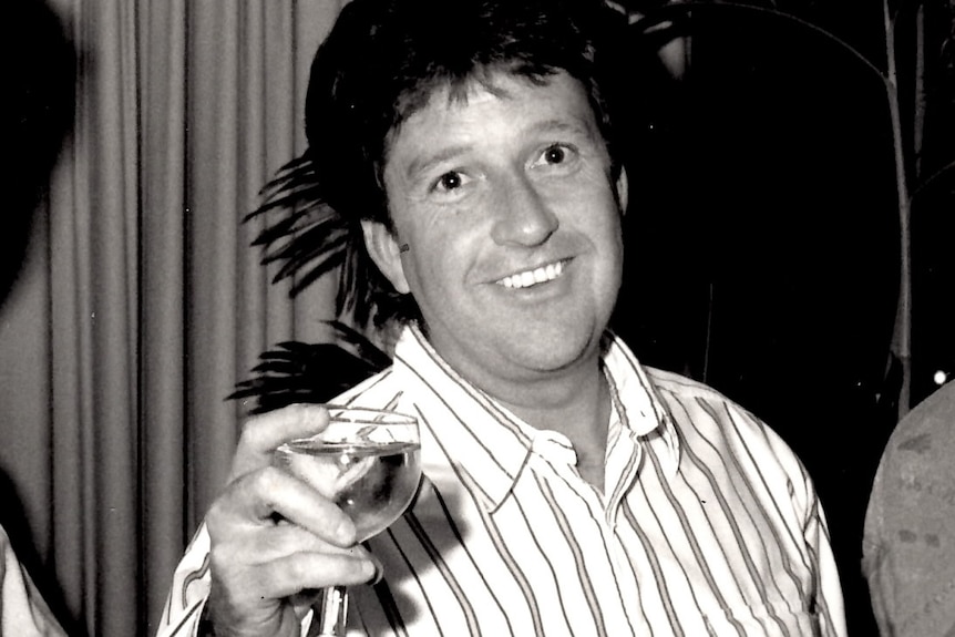 A black and white photo of Denis Handlin. He is toasting with a wine glass while smiling at the camera.