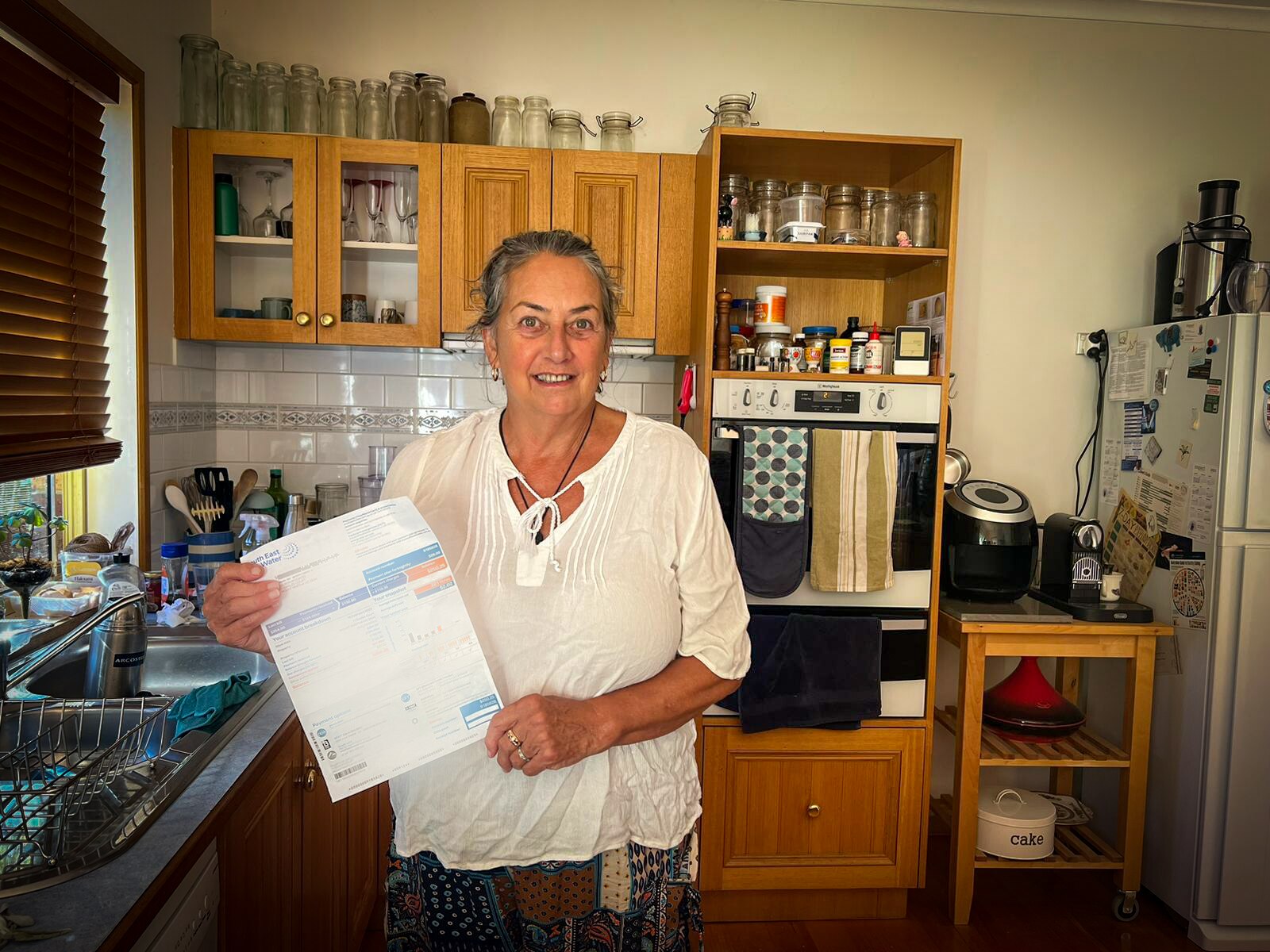 An older woman stands in a kitchen holding a bill