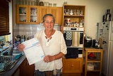 An older woman stands in a kitchen holding a bill