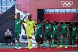 A women's soccer team wearing green uniforms walks out under a red banner gesturing to their heads