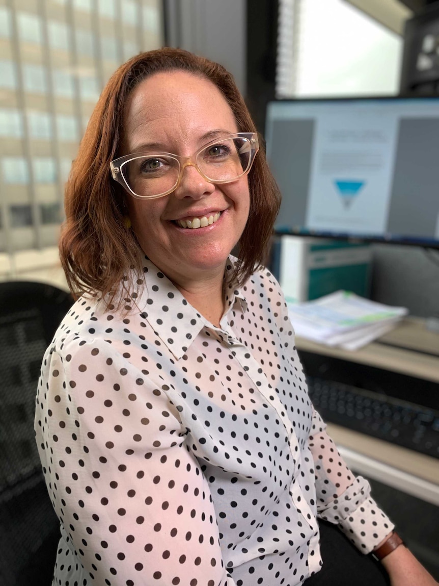 A smiling women wearing glasses sitting in front of a computer screen.