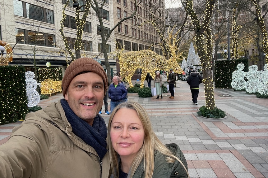 A man and woman stand together for a selfie in front of a display of Christmas lights in trees.