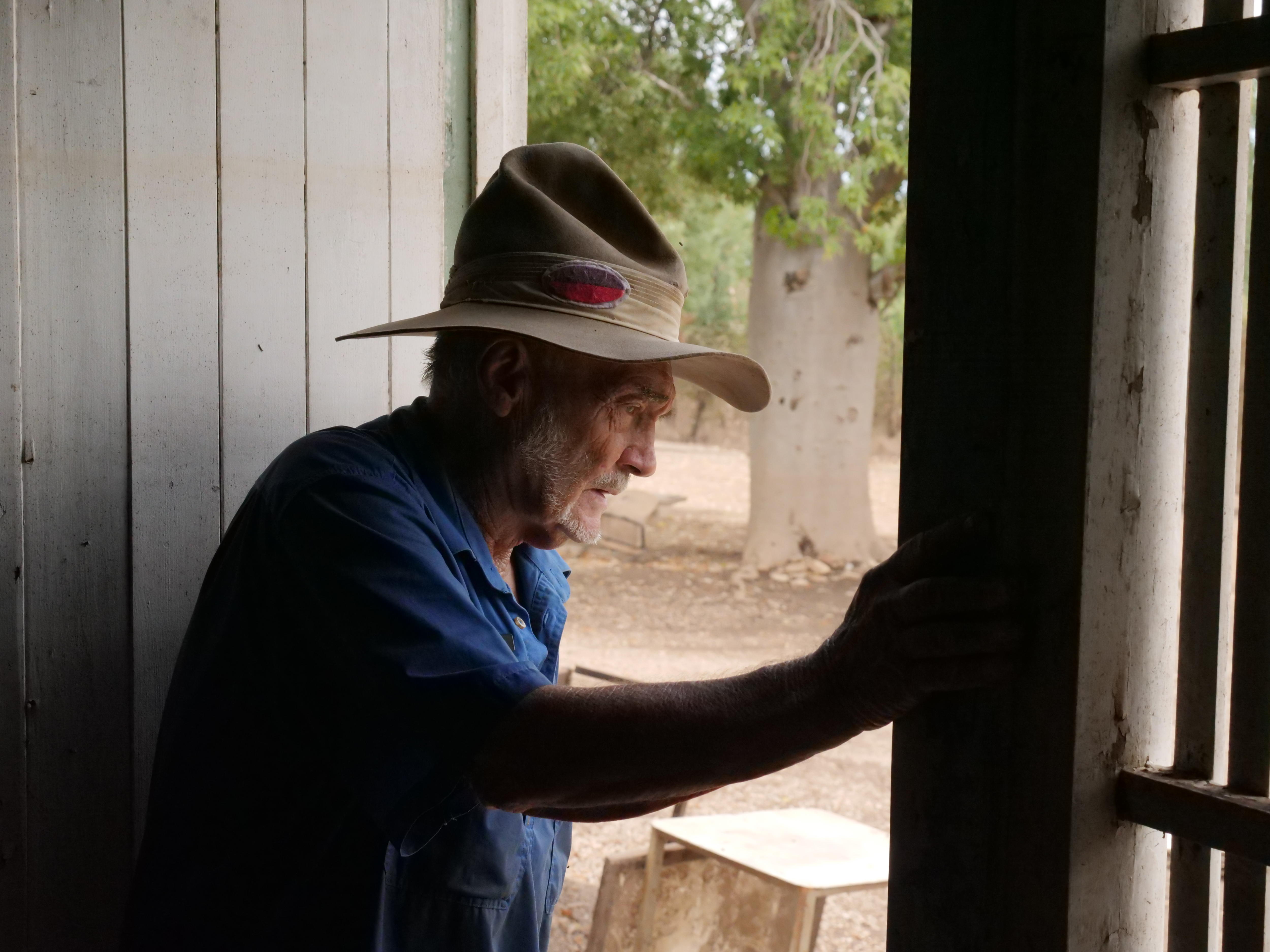 A man in an old-fashioned police hat looks through a doorframe, his face illuminated by the light coming in. 