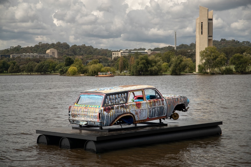 An abandoned Holden painted with designs, mounted on a pontoon on a lake, with an overcast sky and a cityscape behind.