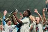 Sydney FC footballer Princess Ibini holding a trophy with her teammate, with their arms raised in victory