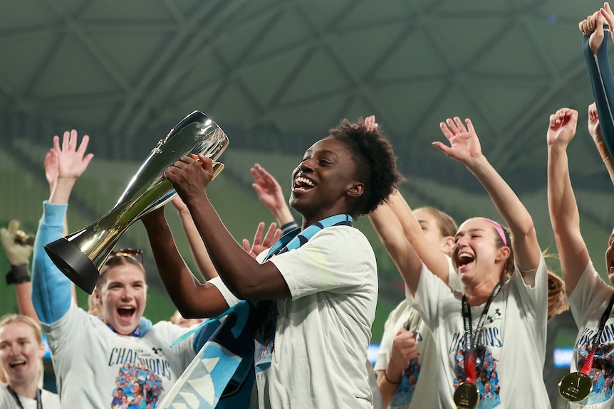 Sydney FC footballer Princess Ibini holding a trophy with her teammate, with their arms raised in victory
