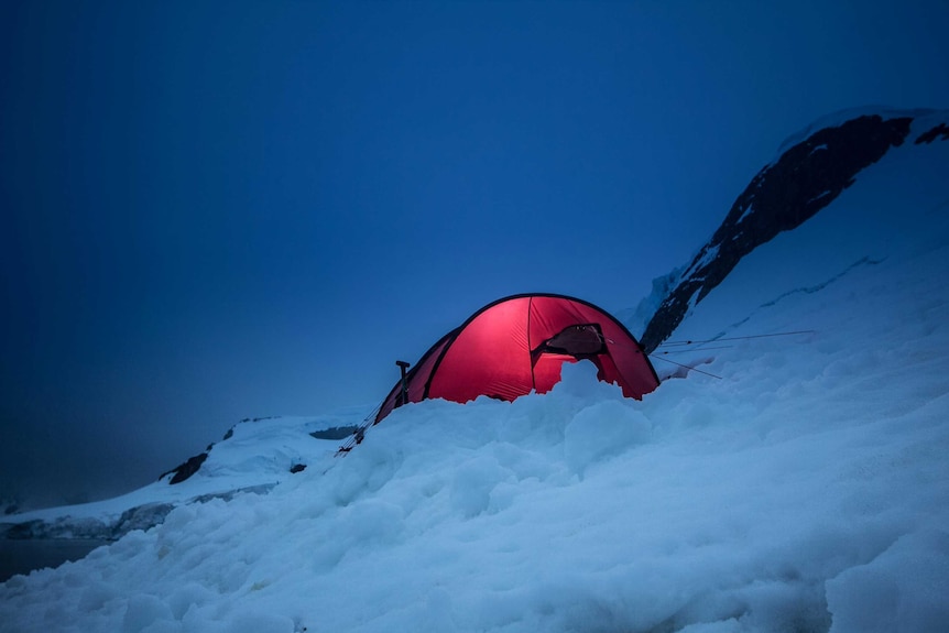 A red tent on the side of an icy slope. It is glowing as there is light inside.