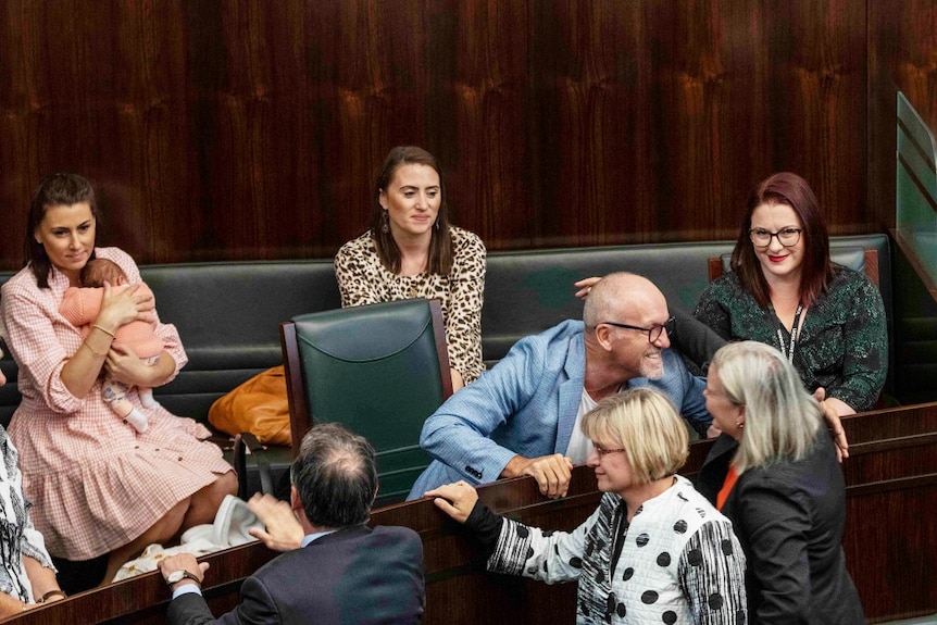 A balding man leans forward to kiss a woman, watched by three women behind him in parliament