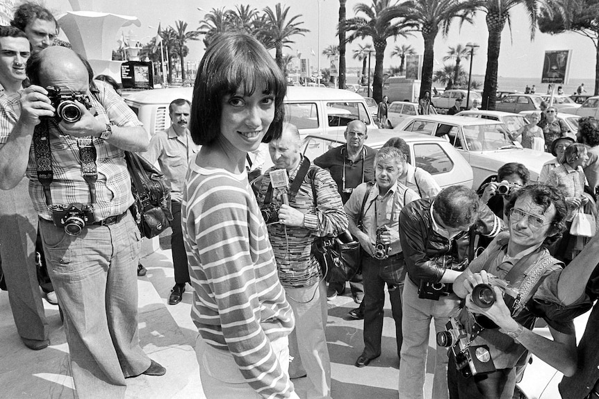 Shelley Duvall stands among a crowd of photographers. She looks over her right shoulder and smiles. 