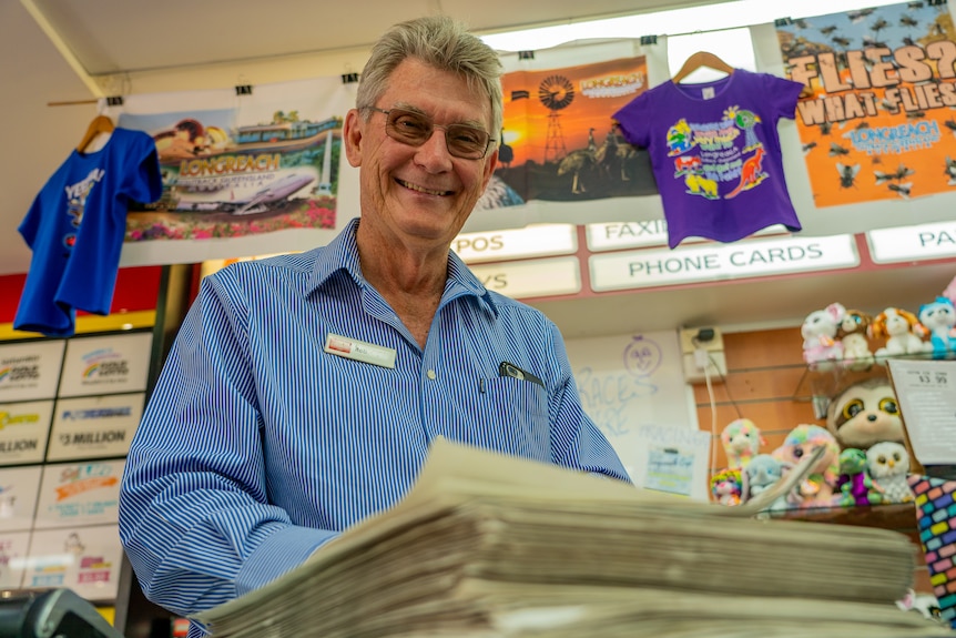 A man standing behind a newsagent counter smiles down at the camera. A stack of newspapers are in the foreground.