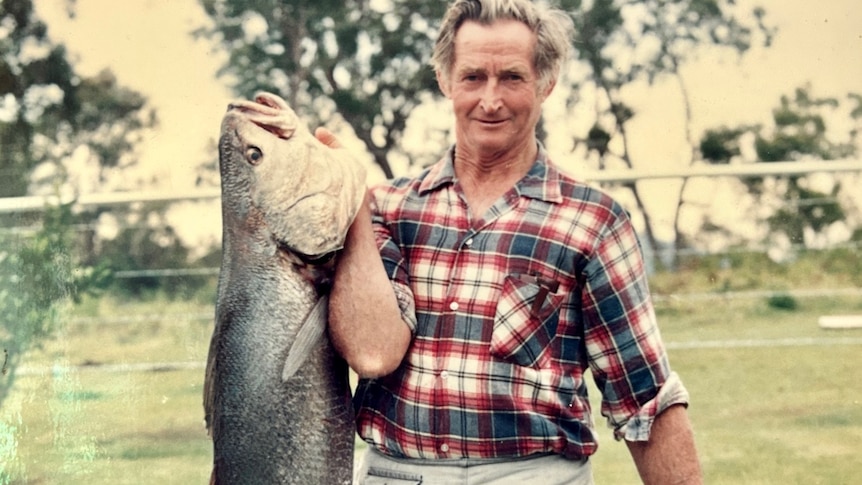 Man in check shirt and light blue shorts holds up a huge fish and smiles.