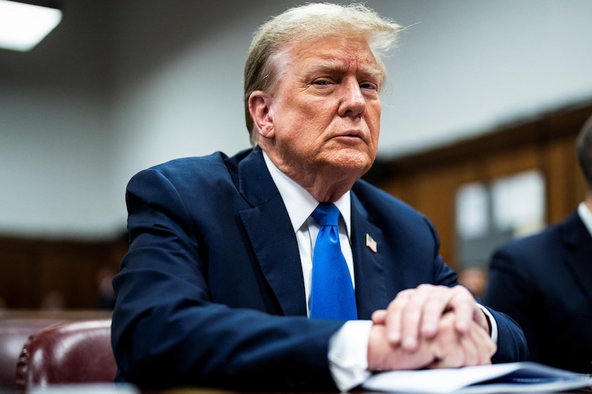 Donald Trump takım elbiseli, mavi kravatlı ve beyaz gömlekli bir masanın arkasında oturuyor ve sert bir şekilde kameraya bakıyor