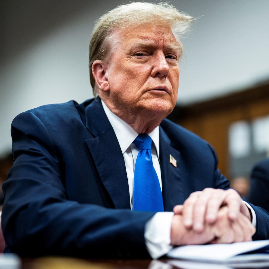 Donald Trump sits in a suit, blue tie and white shirt looking sternly at the camera behind a desk