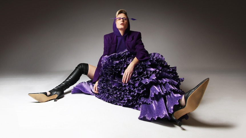 Róisín Murphy seated, wearing knee high heels and a purple dress with hood, balancing an arrow on her nose