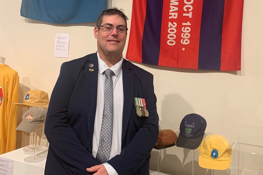 A tall man wearing glasses and a suit with military medals on his left breast, standing with an INTERFET flag.